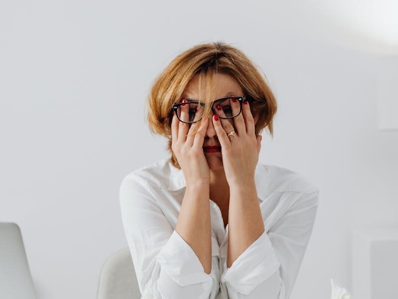 A stressed woman in eyeglasses and white shirt sits at her desk, overwhelmed and exhausted.