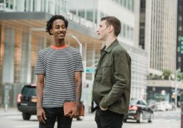 Smiling multiracial friends in casual clothes standing on street near roadway and talking against buildings in city in daytime