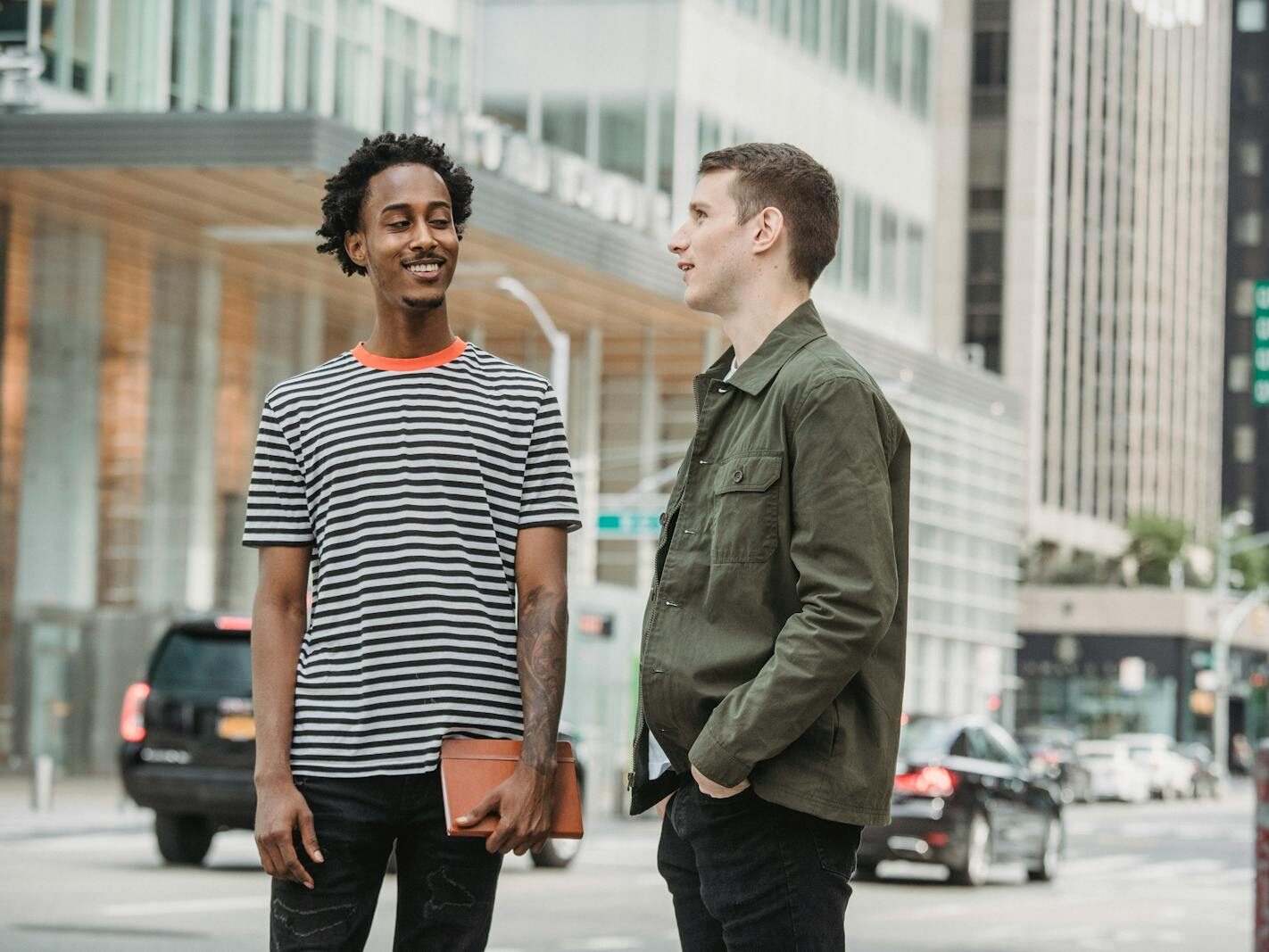 Smiling multiracial friends in casual clothes standing on street near roadway and talking against buildings in city in daytime