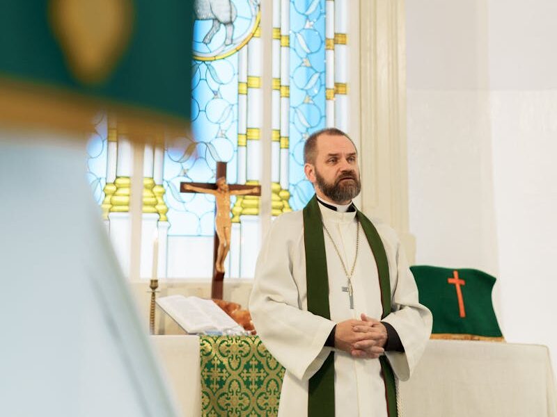 Pastor in cassock leads prayer in a historic church with stained glass windows and altar.