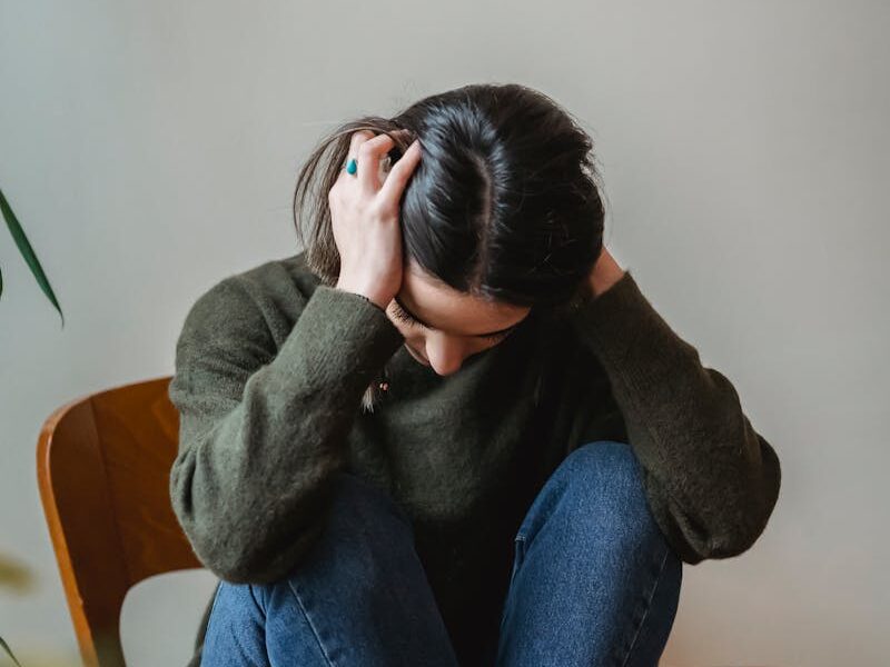 A young woman sitting on a chair indoors, holding her head in distress.