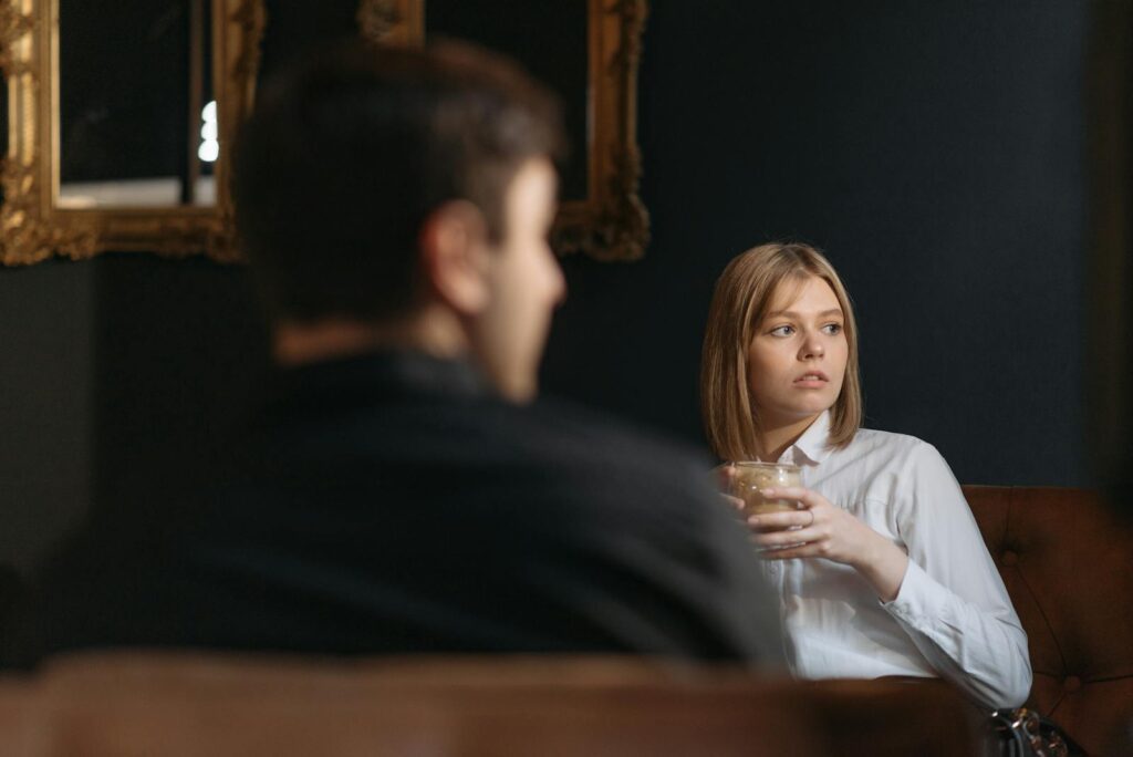 A woman sits thoughtfully in a cafe holding a drink, looking away with an introspective expression.