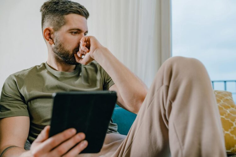 A man in casual attire, sitting indoors, holding a tablet and looking thoughtful.