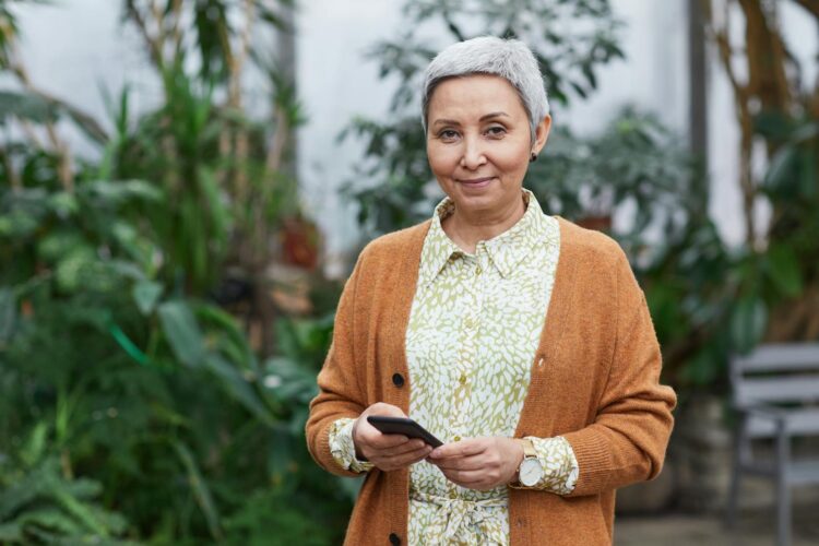 Elderly woman with smartphone standing in a greenhouse, smiling.