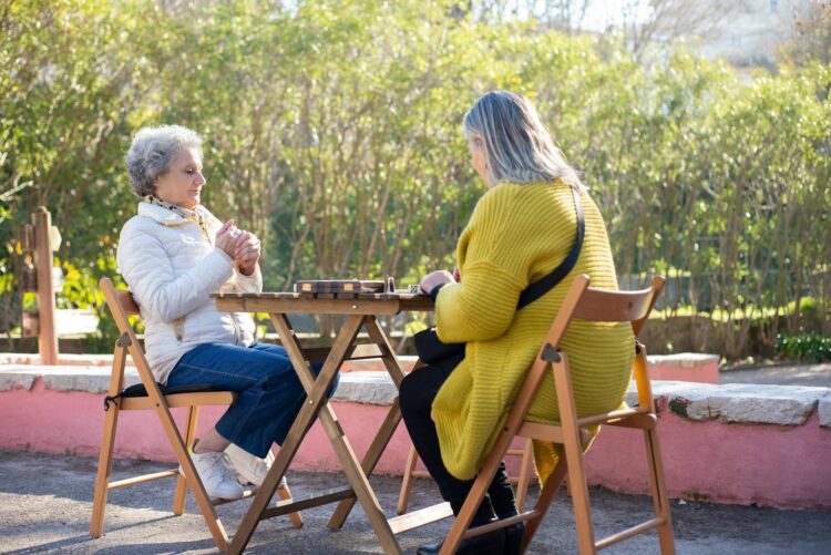 Senior women enjoying a game of dominoes at a wooden table in a sunny park setting.