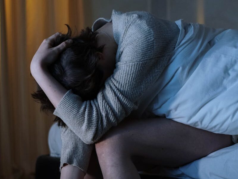A woman sits on her bed, experiencing anxiety and insomnia in a dimly lit room.