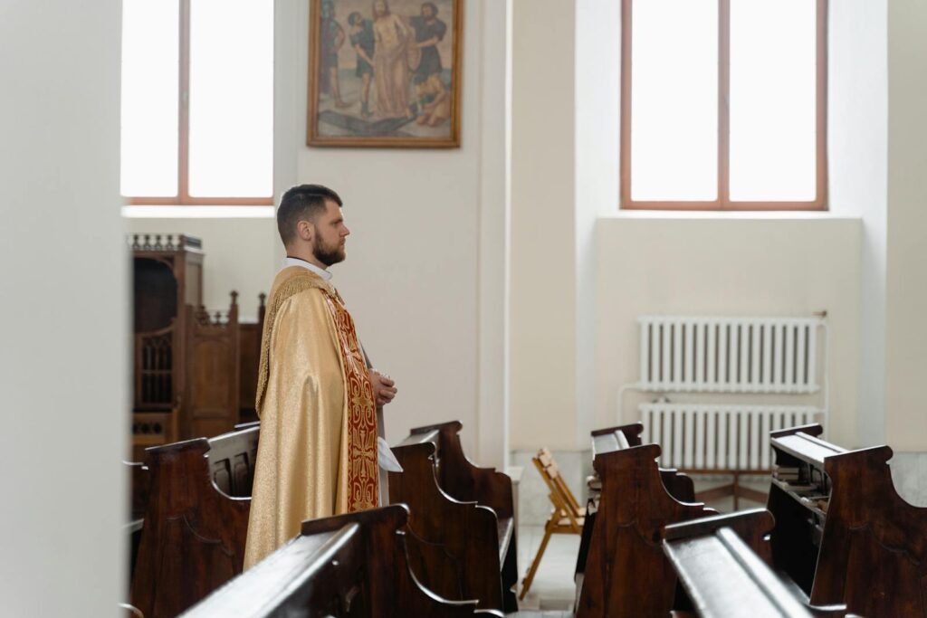 A priest in religious vestments stands inside a serene church with classic wooden pews and religious artwork.