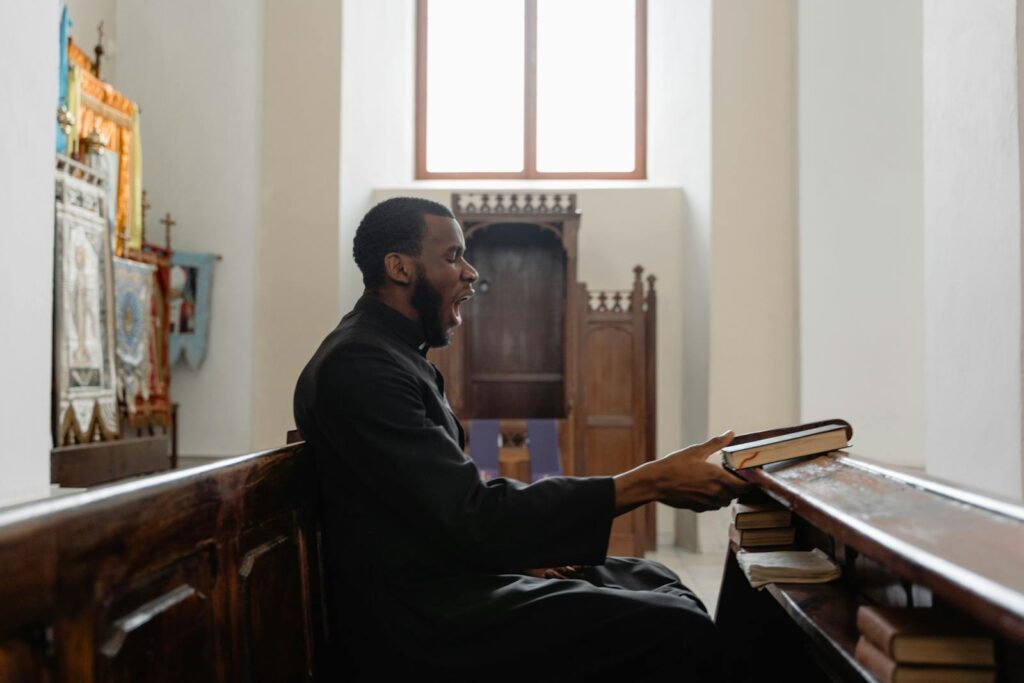 A tired priest yawns while sitting in a church pew wearing a cassock.