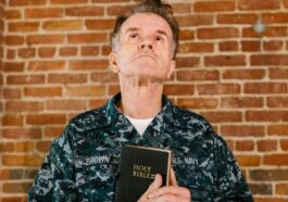 A senior US Navy veteran stands against a brick wall holding a Holy Bible.