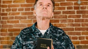 A senior US Navy veteran stands against a brick wall holding a Holy Bible.