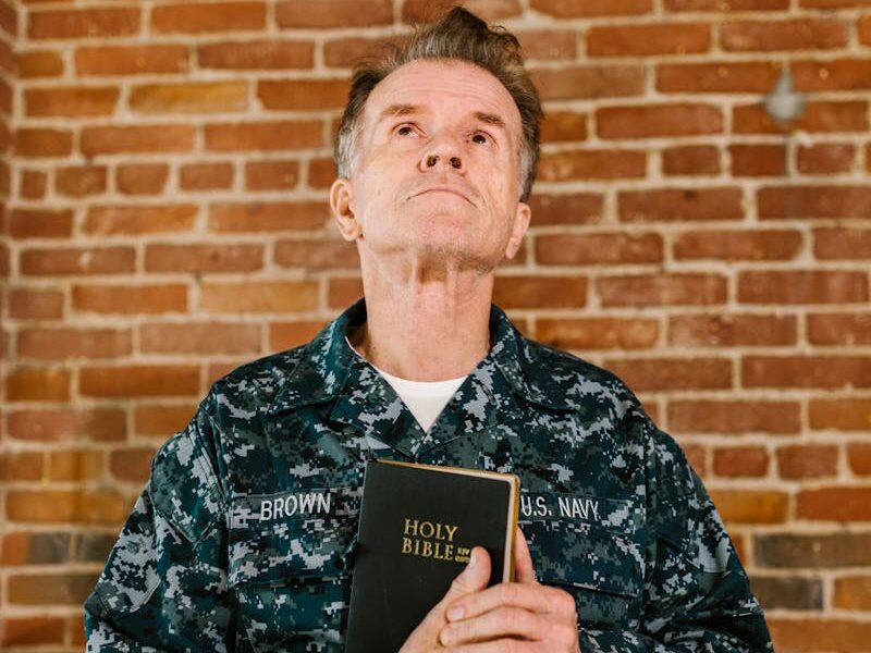 A senior US Navy veteran stands against a brick wall holding a Holy Bible.