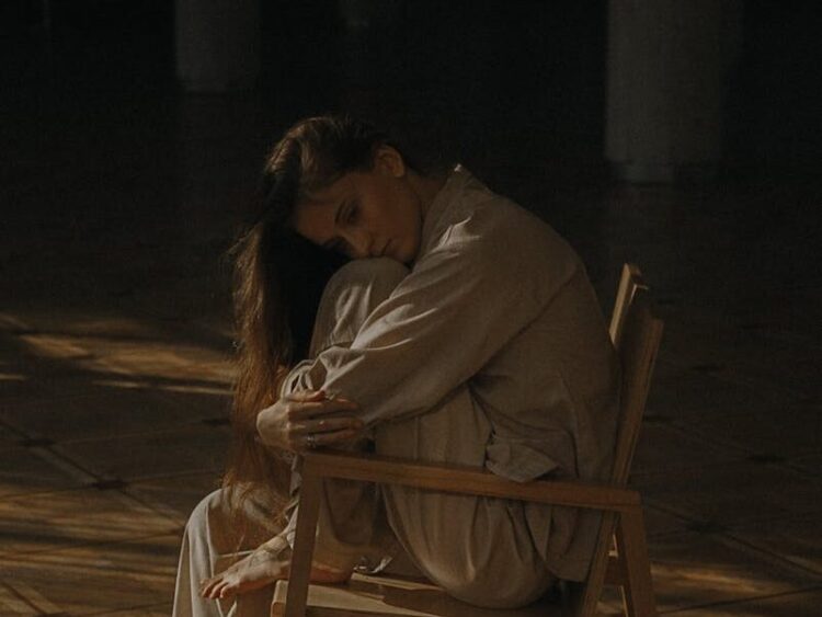 A woman in casual attire sits alone on a chair in a dimly lit room, expressing deep emotions.