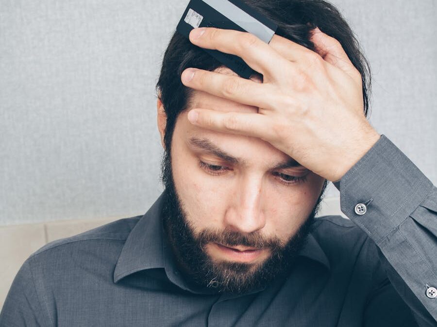 A stressed man looks at his smartphone, holding a credit card in his hand.