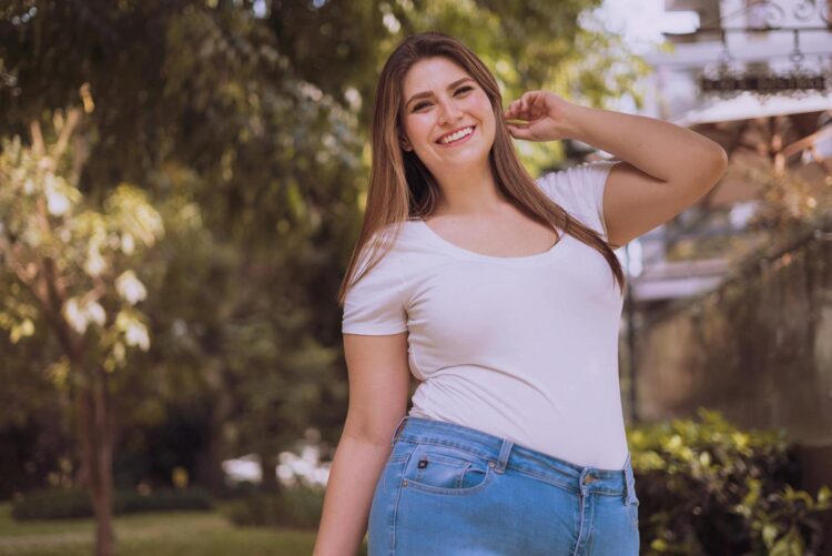 A cheerful plus-size Hispanic woman posing confidently in a serene outdoor setting.