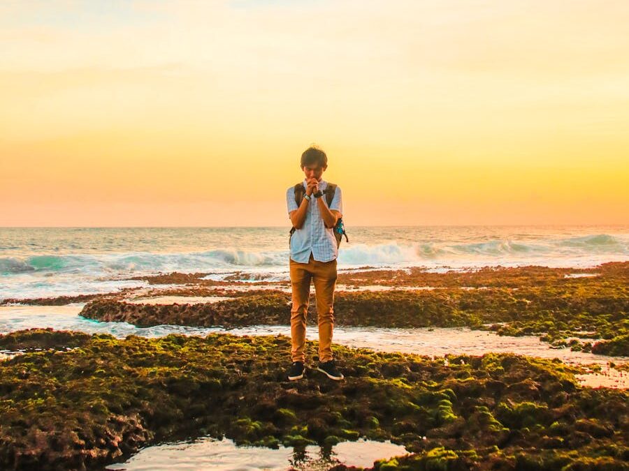 A man stands on mossy rocks by the ocean, praying during a vibrant sunset, creating a serene and introspective scene.
