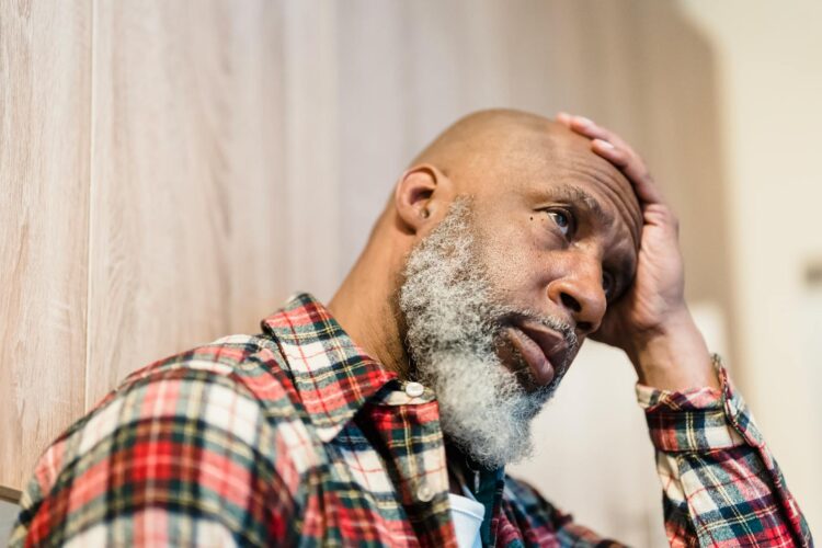Close-up of a thoughtful bald man with a beard, sitting indoors with his hand on his head.