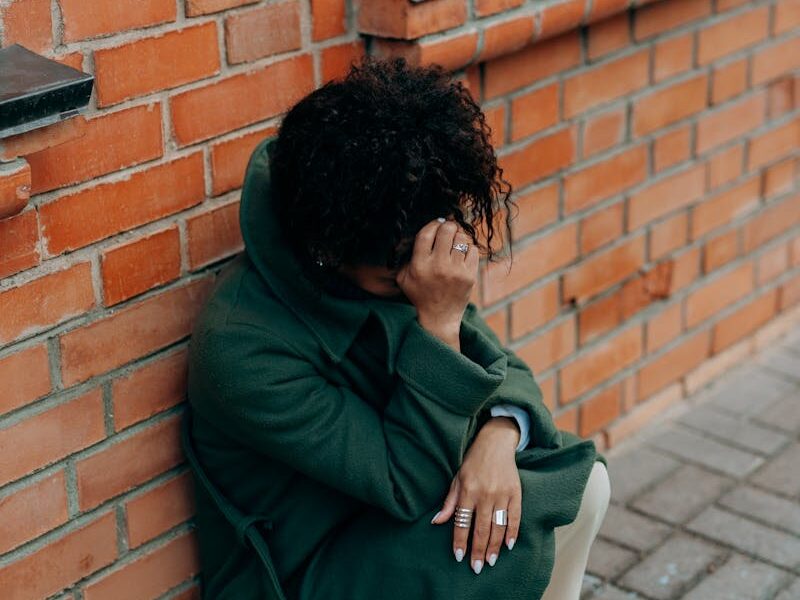 A woman in a green coat sits against a brick wall, deep in thought.