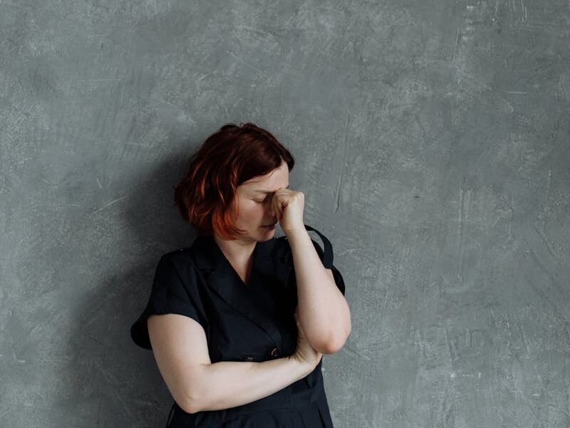 Pensive woman in black dress leans against textured gray wall, deep in thought.