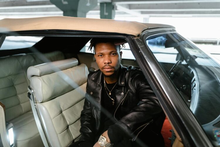 African American man in leather jacket seated in vintage convertible car.