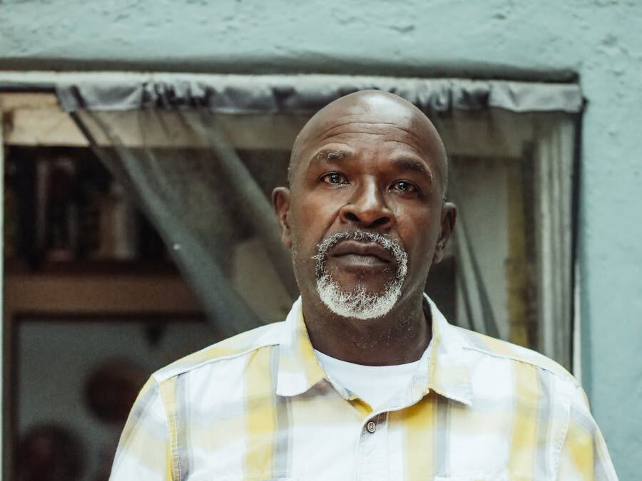 A senior man stands indoors wearing a checkered shirt, appearing contemplative.