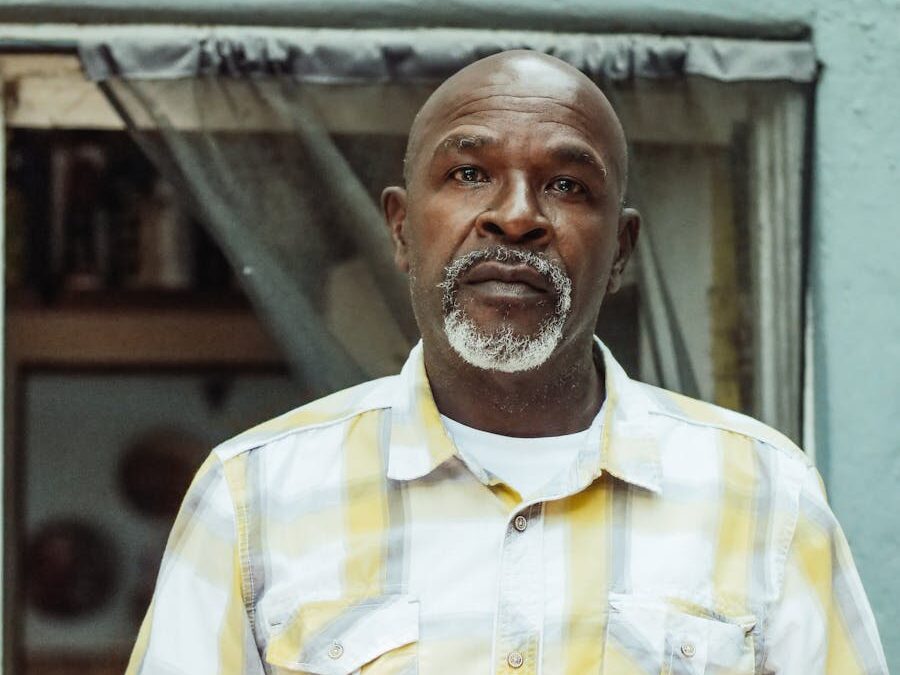 A senior man stands indoors wearing a checkered shirt, appearing contemplative.