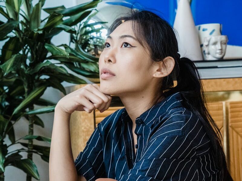 Asian woman sitting thoughtfully indoors beside indoor plants.