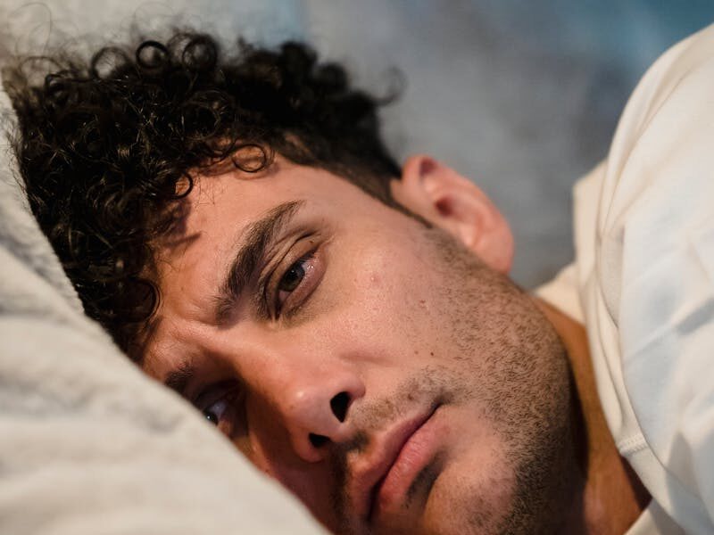 A man lies in bed, deep in thought with a pensive expression, during a nighttime moment.