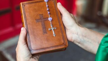 Hands holding a leather-bound Bible with rosary beads, in front of a red door, symbolizing faith.