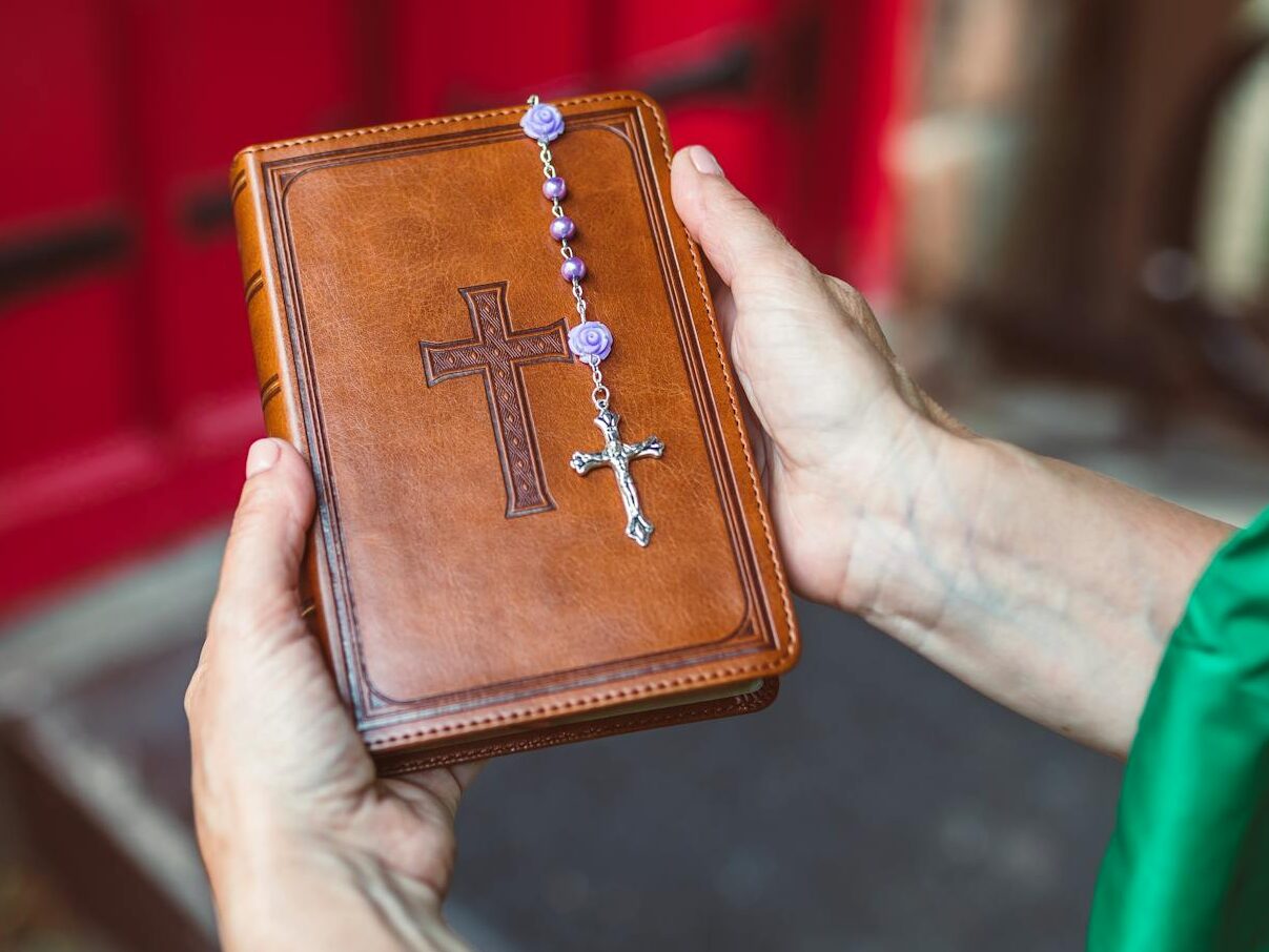 Hands holding a leather-bound Bible with rosary beads, in front of a red door, symbolizing faith.