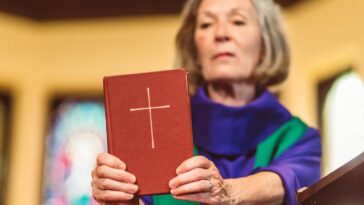 An elderly female priest holding a Bible inside a church setting.