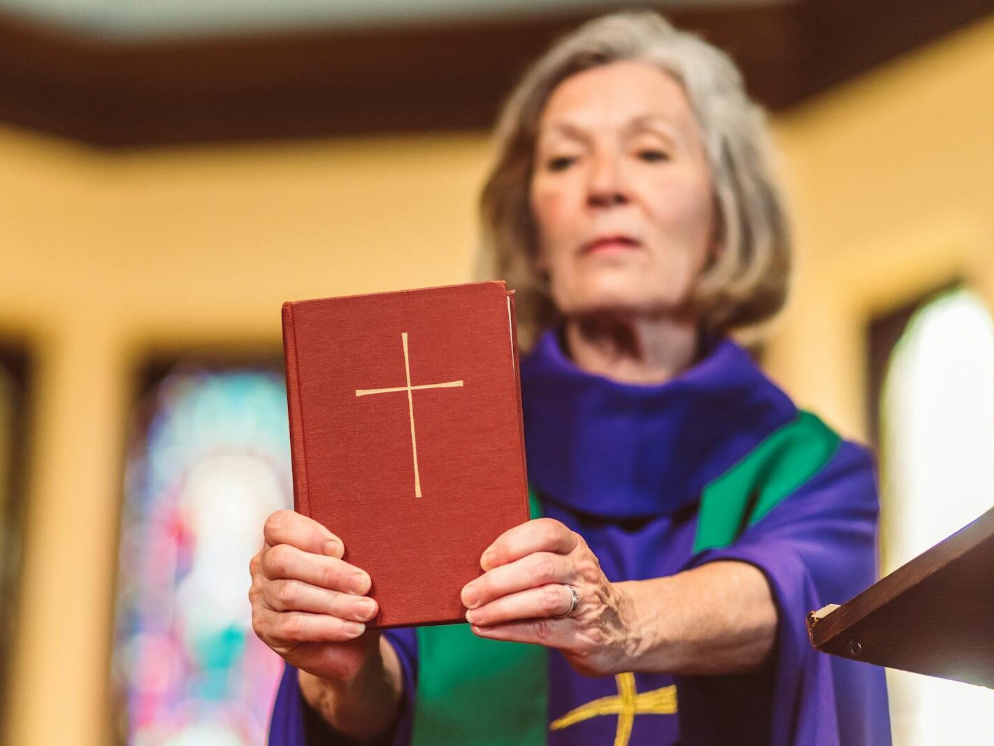 An elderly female priest holding a Bible inside a church setting.