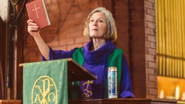 Elderly female priest delivering a sermon in a traditional church setting, holding a Bible.