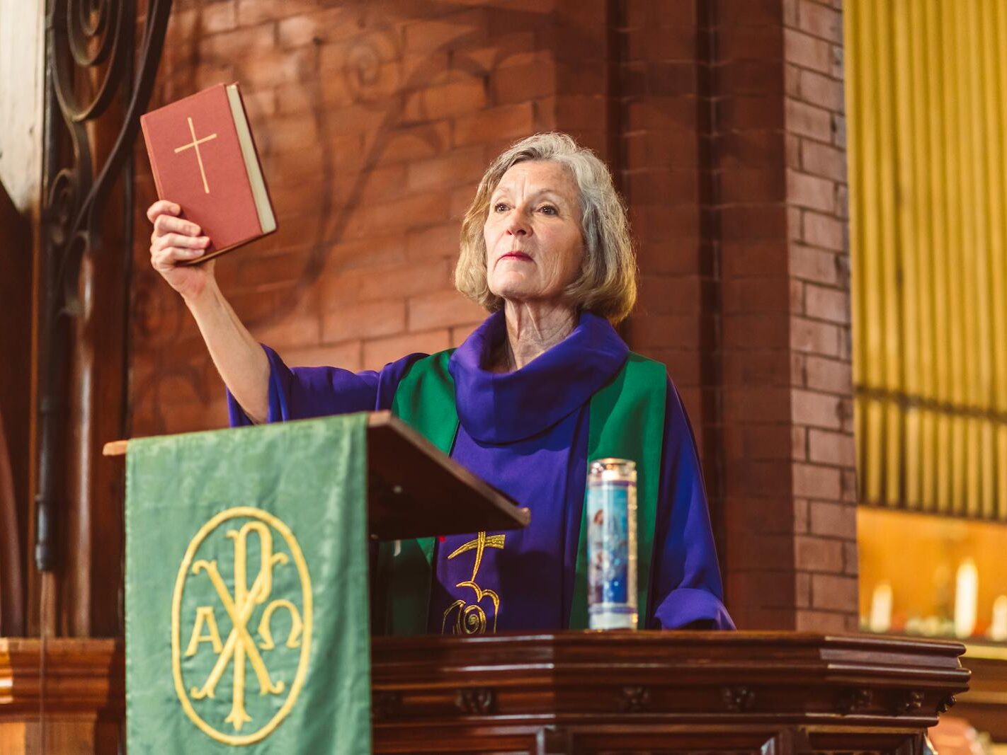 Elderly female priest delivering a sermon in a traditional church setting, holding a Bible.