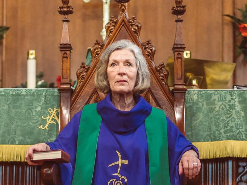 Elderly female priest in traditional blue vestments sitting on a church throne, holding a book.