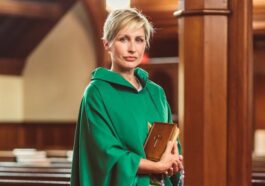 A woman priest stands in a church in traditional green chasuble, holding a Bible.