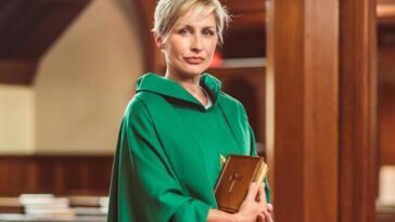 A woman priest stands in a church in traditional green chasuble, holding a Bible.