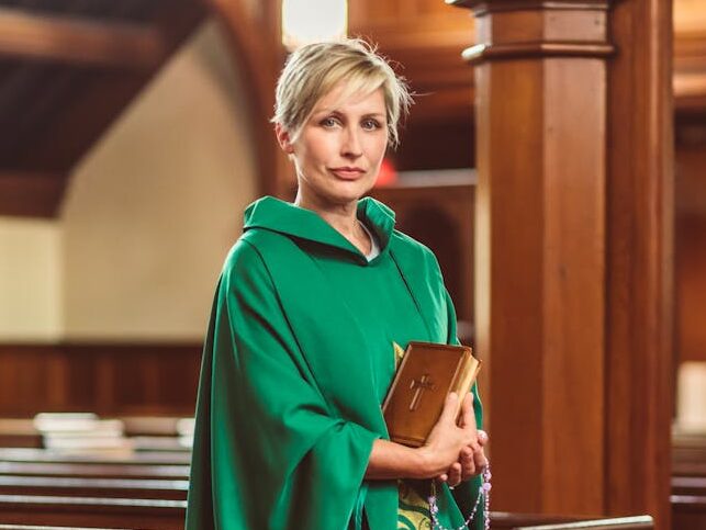 A woman priest stands in a church in traditional green chasuble, holding a Bible.