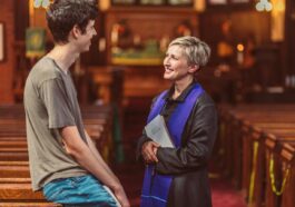 A priest holds a Bible while conversing with a young man in a warm, light-filled church interior.