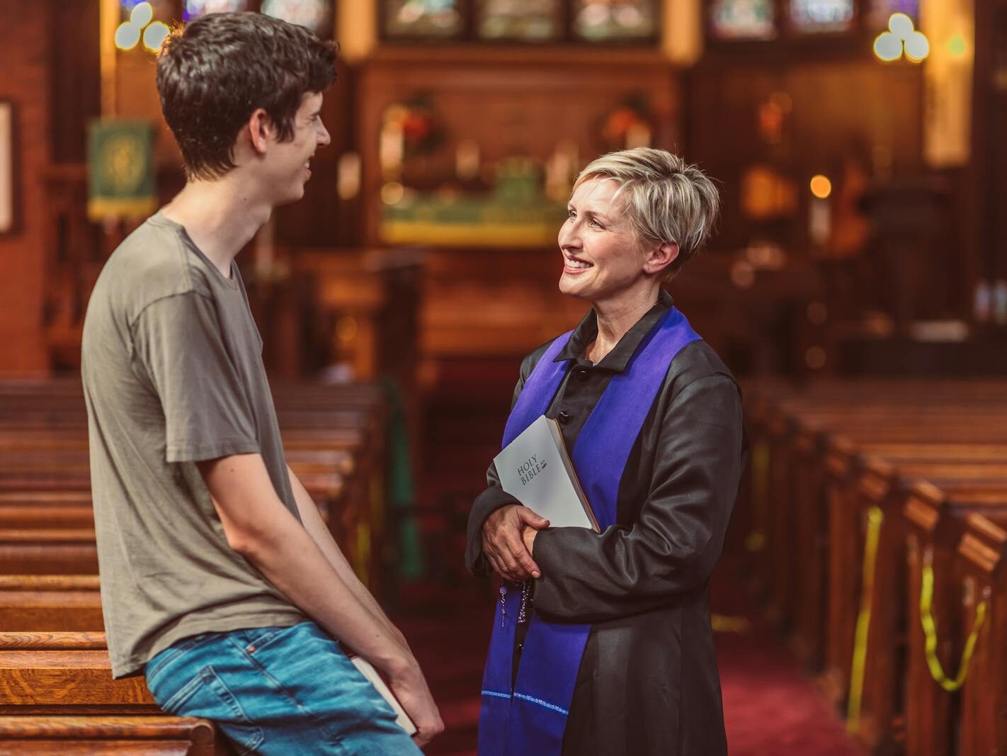 A priest holds a Bible while conversing with a young man in a warm, light-filled church interior.