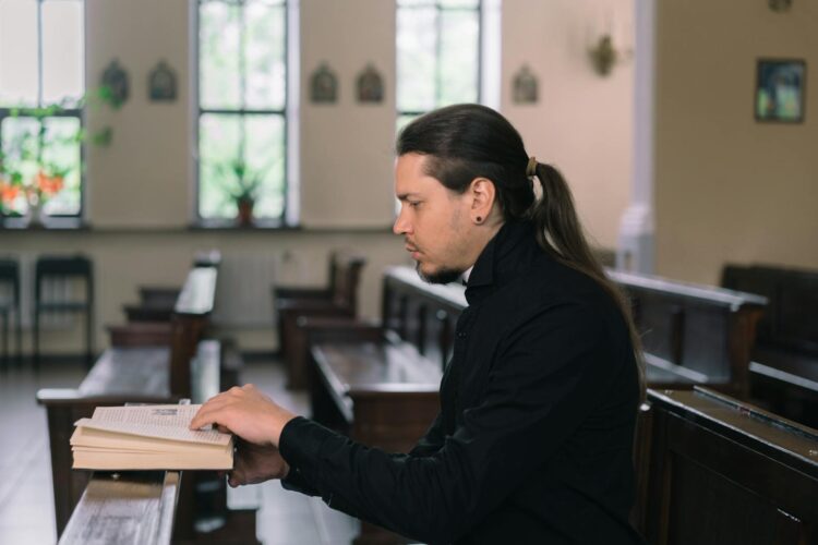 A man with long hair reading a book in a church pew, highlighting faith.