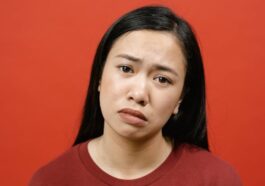 A studio shot of an Asian woman in a red shirt against a red background, showing a sad expression.