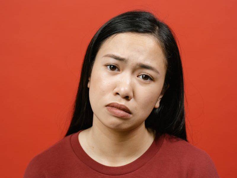 A studio shot of an Asian woman in a red shirt against a red background, showing a sad expression.