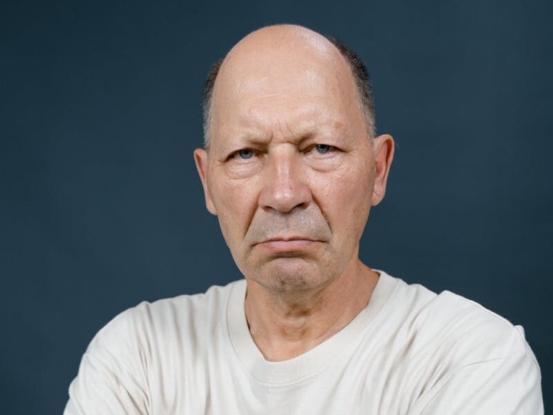 Portrait of a bald elderly man with arms crossed showing intense expression in a studio setting.