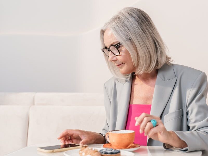 Senior woman in a gray blazer enjoying coffee and pastries at a table with a phone.