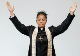 A female priest in black robe praying with arms raised and eyes closed against a white studio background.