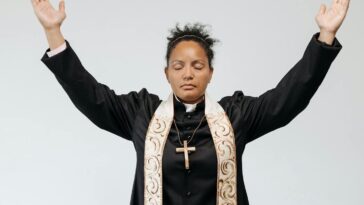 A female priest in black robe praying with arms raised and eyes closed against a white studio background.