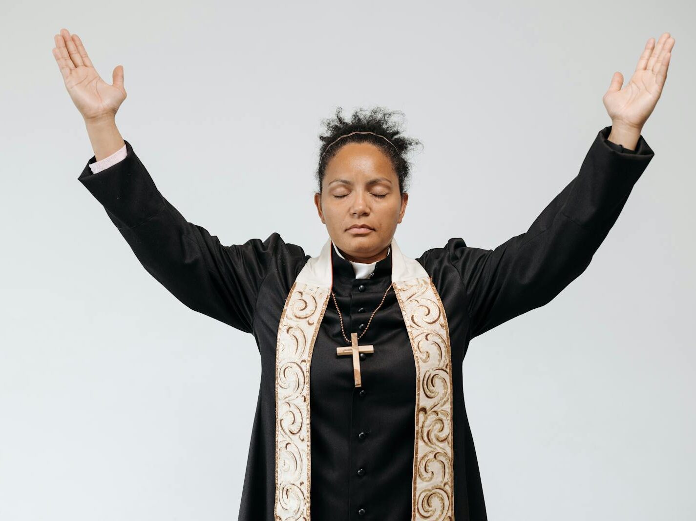 A female priest in black robe praying with arms raised and eyes closed against a white studio background.