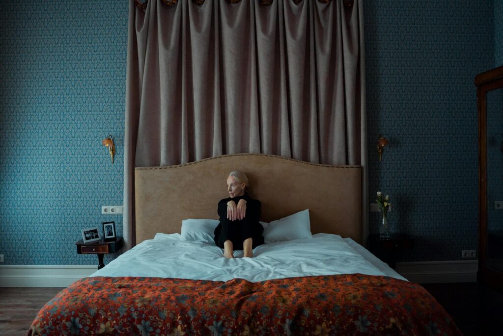 Elderly woman sitting on a bed, contemplating in a cozy and luxurious room.