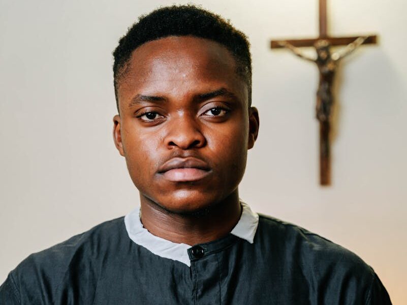 Portrait of a young man in a black robe holding a Bible in a church setting with a crucifix in the background.