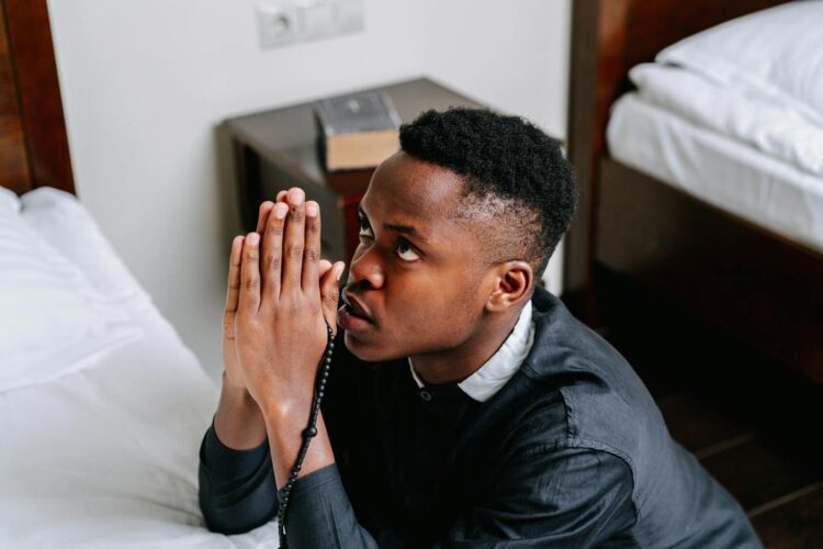 Young man kneeling in prayer with rosary beads in a peaceful room setting.
